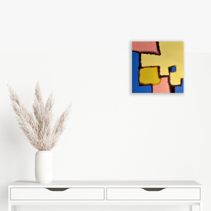 The artwork 'Thawed Geometry' features abstract geometric shapes in vibrant yellow, coral pink, and electric blue, separated by bold black lines creating a modernist composition. The shapes appear to interlock like puzzle pieces on a square canvas, finished with synthetic glazes. 24x24 inches. By Sabrina Puppin.